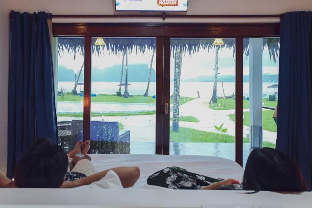 Una habitación con una cama, dos personas acostadas y un ventanal con vista a una playa y palmeras.