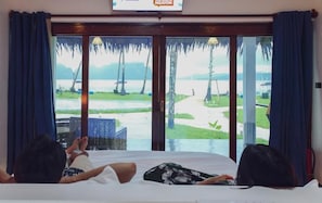 A room with a bed, two people lying down, and a large window with a view of a beach and palm trees.