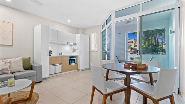Una cocina moderna con armarios blancos, un comedor con una mesa redonda y sillas blancas, y un balcón con vistas a palmeras.