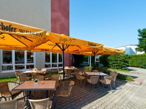 Ein Café mit Außenbereich, Tische und Stühle unter gelben Sonnenschirmen, ein Gebäude mit der Aufschrift 'Weizen' und ein klarer Himmel.