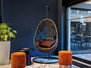 Ein Hängekorbstuhl mit Kissen, ein runder, orangefarbener Hocker und ein Blumentopf in einem modernen Interieur.