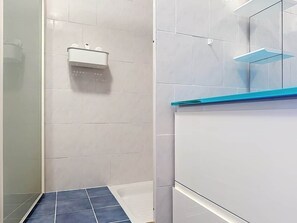 Ein Badezimmer mit weiß gefliesten Wänden, einem blauen Fliesenboden in der Dusche, einem weißen Waschbecken mit Seifenspender und einer Glasablage an der Wand.
