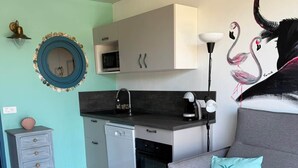 Een kleine leefruimte met een kitchenette, een grijze bank met een turquoise kussen en een muurschildering van flamingo's.