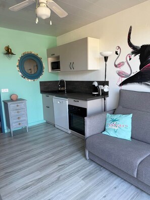 Ein kompakter Wohnraum mit einer Küchenzeile, einem grauen Sofa mit einem türkisfarbenen Kissen und einer Wandmalerei von Flamingos.