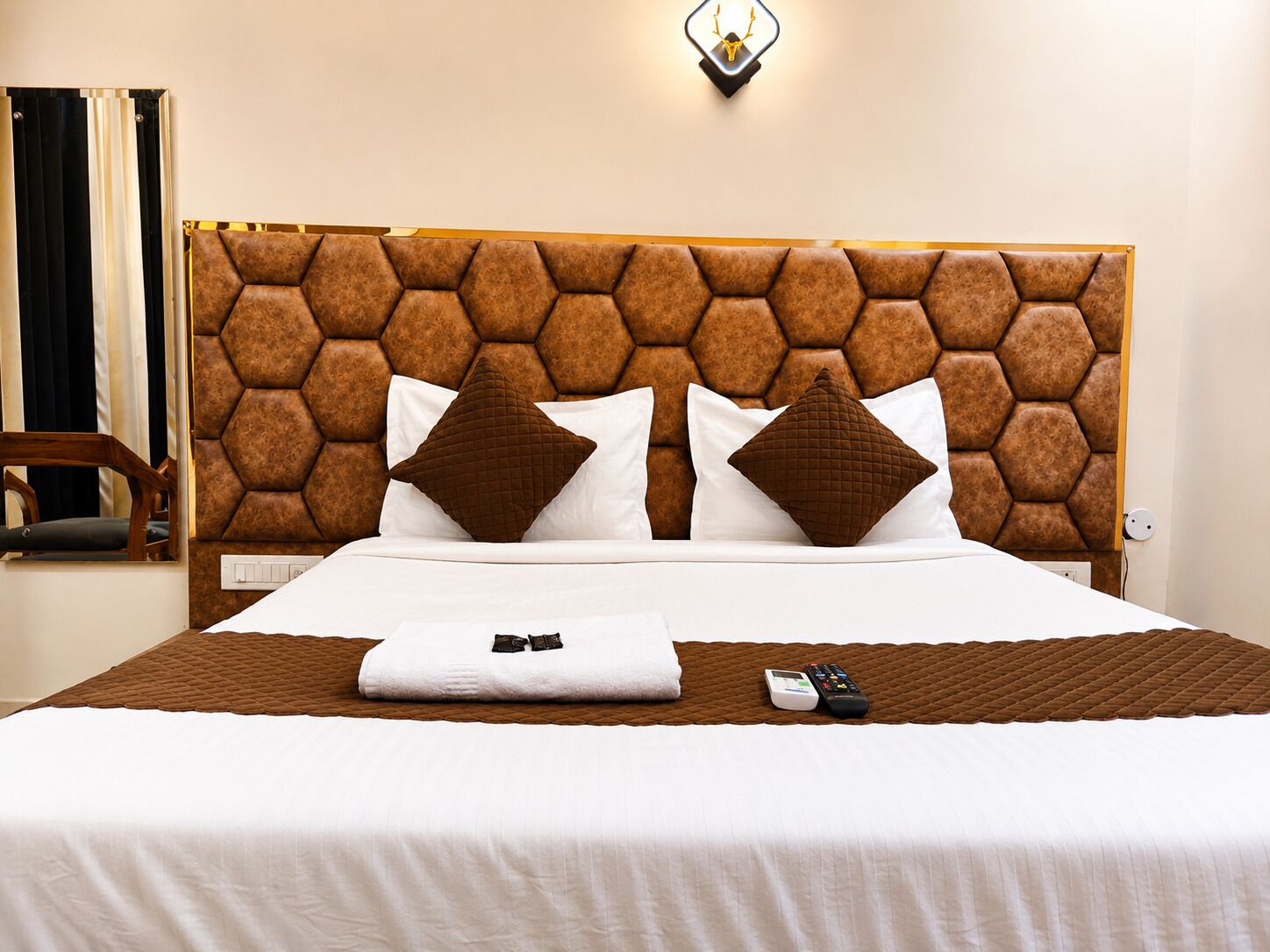 A hotel room with a hexagonal headboard, a bed with white linens, two brown pillows, a remote control, and a folded towel on the bed.