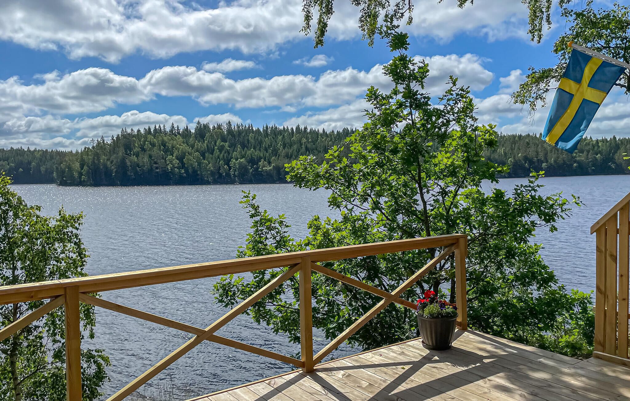 A wooden deck with a railing overlooking a lake, surrounded by trees and a flag.