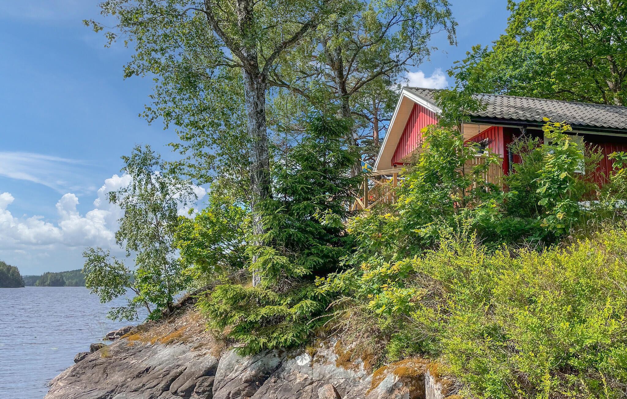 A lakeside cabin with a red exterior, surrounded by dense greenery and rocky terrain.