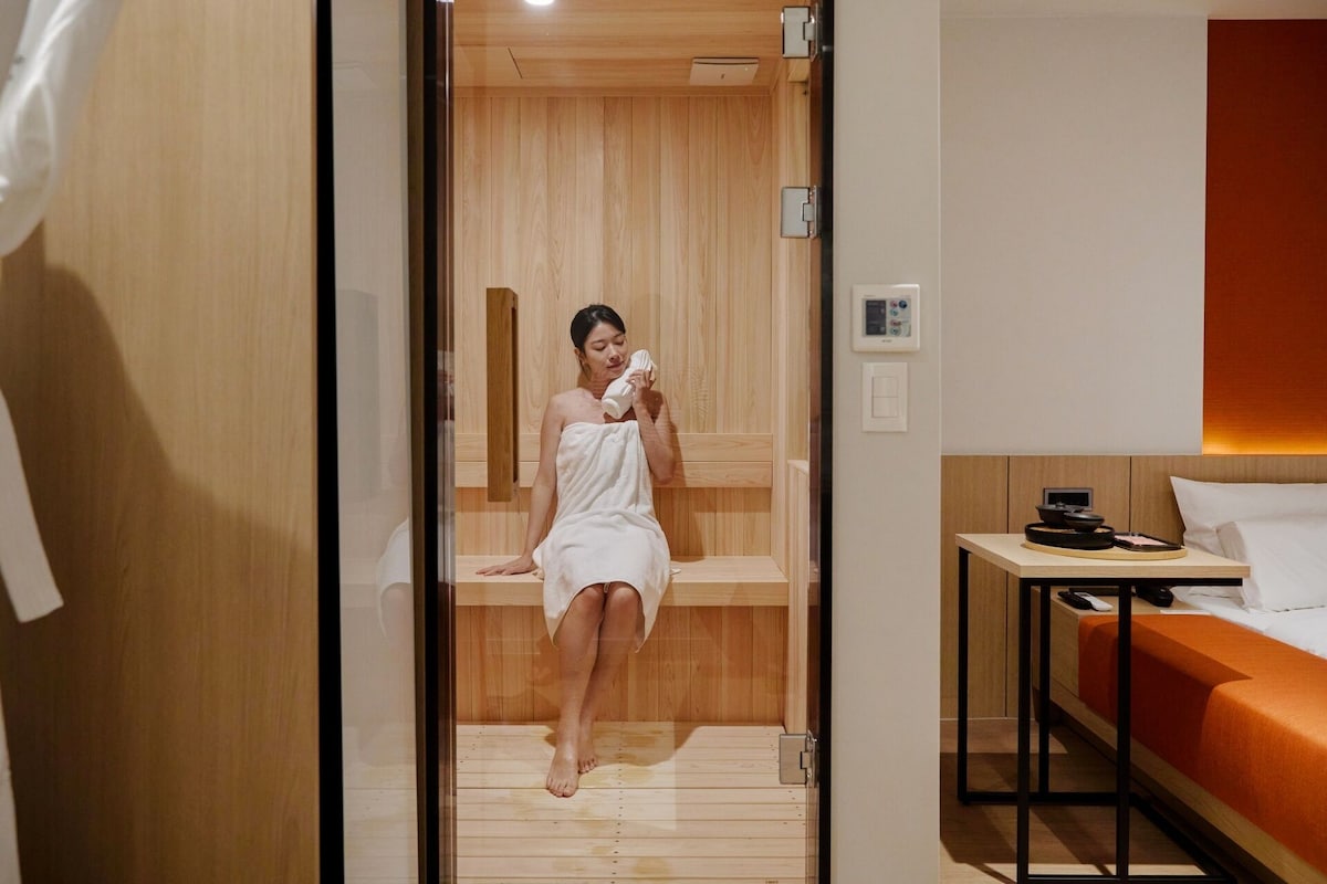 Uma pessoa de toalha num sauna de madeira com uma cama e uma mesa com um telemóvel e uma tigela.