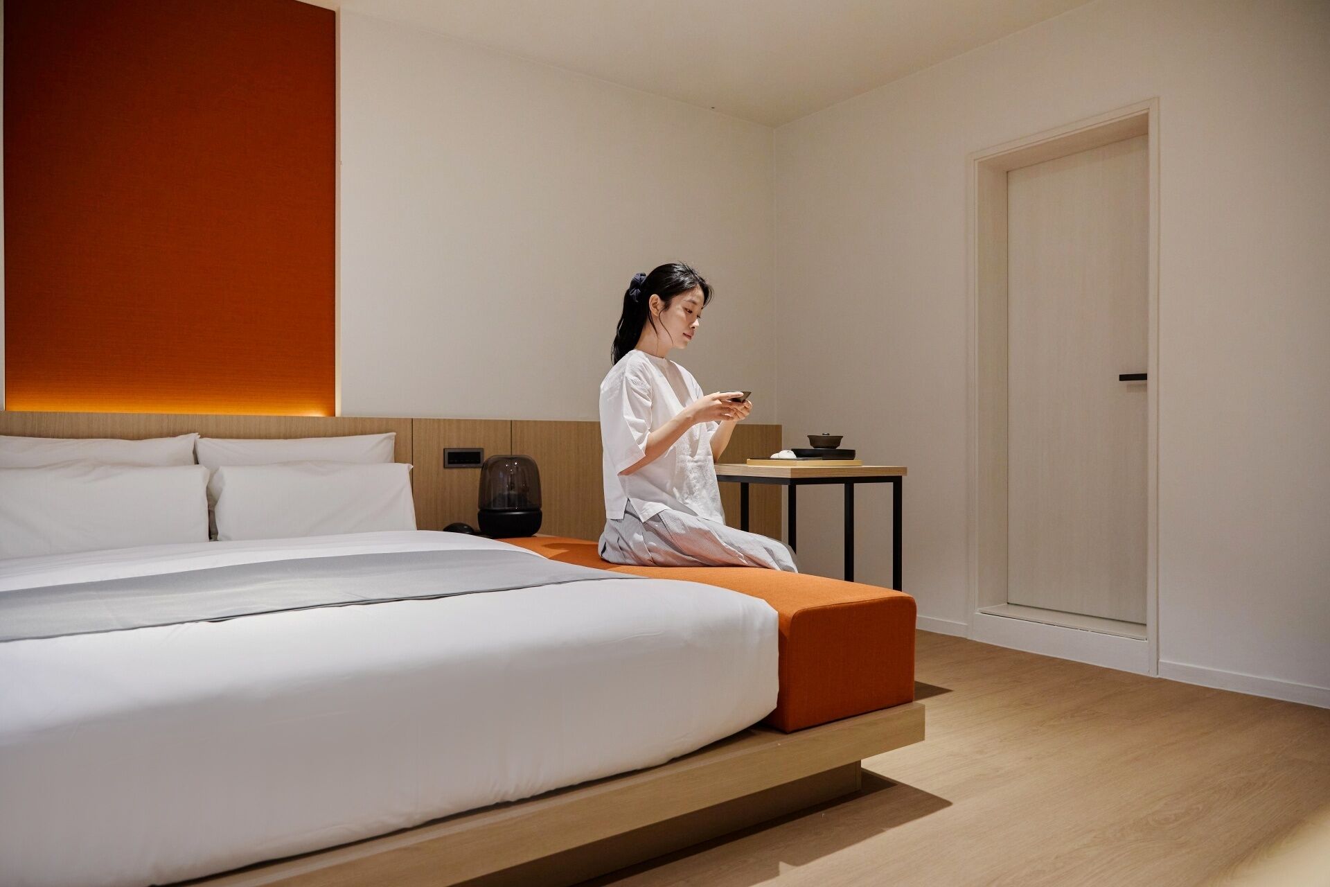Ett modernt hotellrum med en säng, ett litet bord med en skål på, och en person som sitter på sängen.