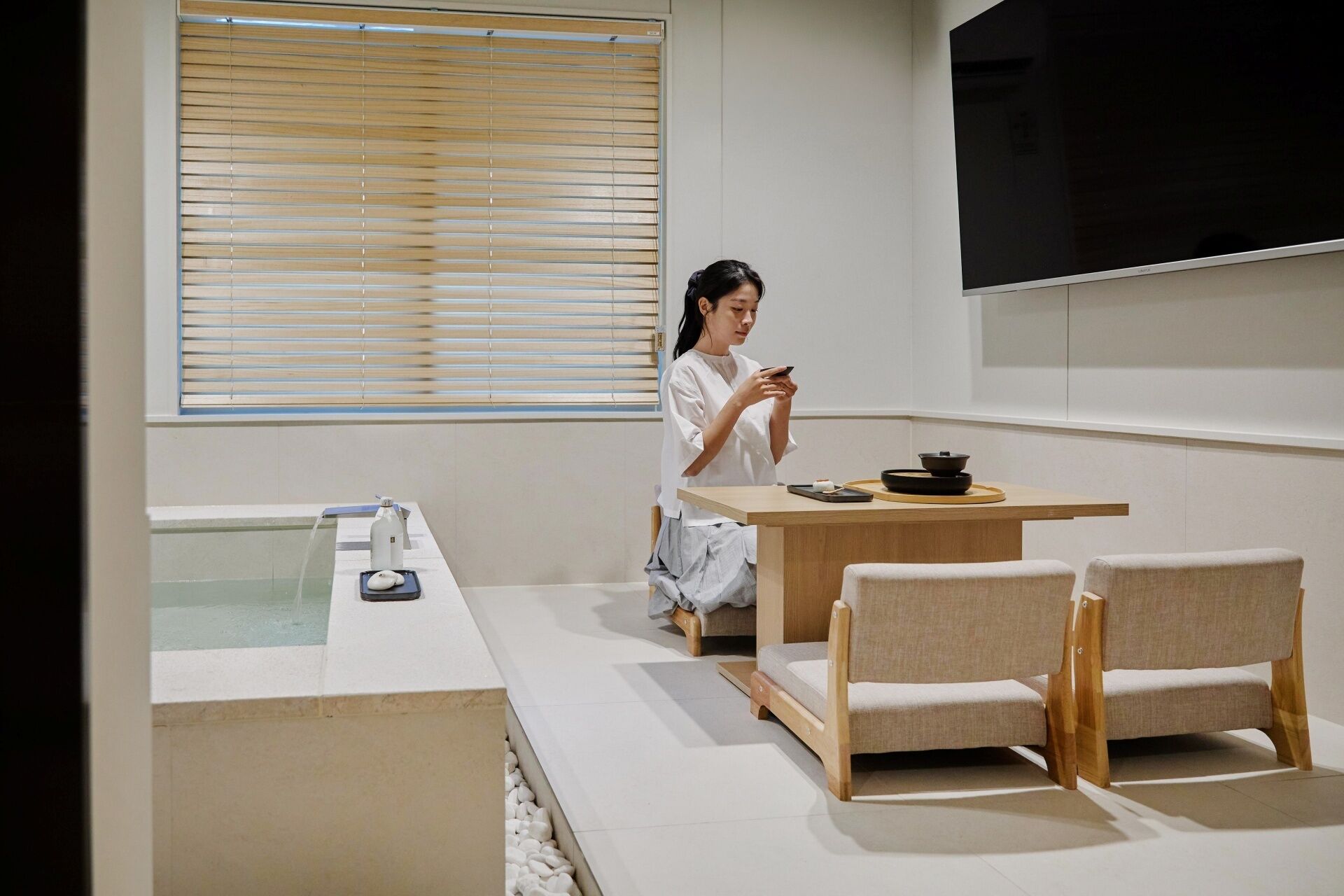 A modern room with a wooden table, two chairs, a TV mounted on the wall, and a window with blinds.