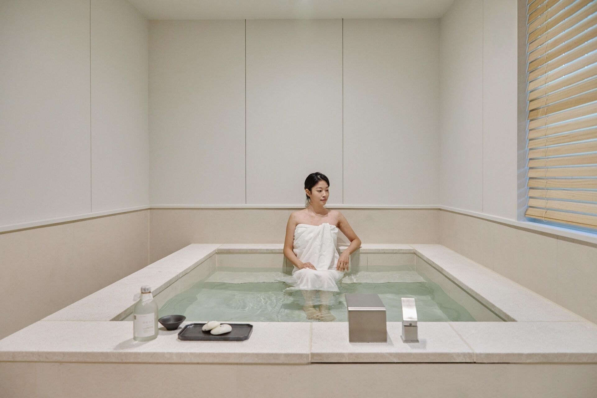 A person in a bathtub with a towel, a bottle, and a tray with items on the edge.