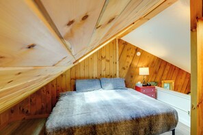 Um quarto com teto inclinado de madeira, uma cama com manta cinzenta e uma mesa de cabeceira com abajur.