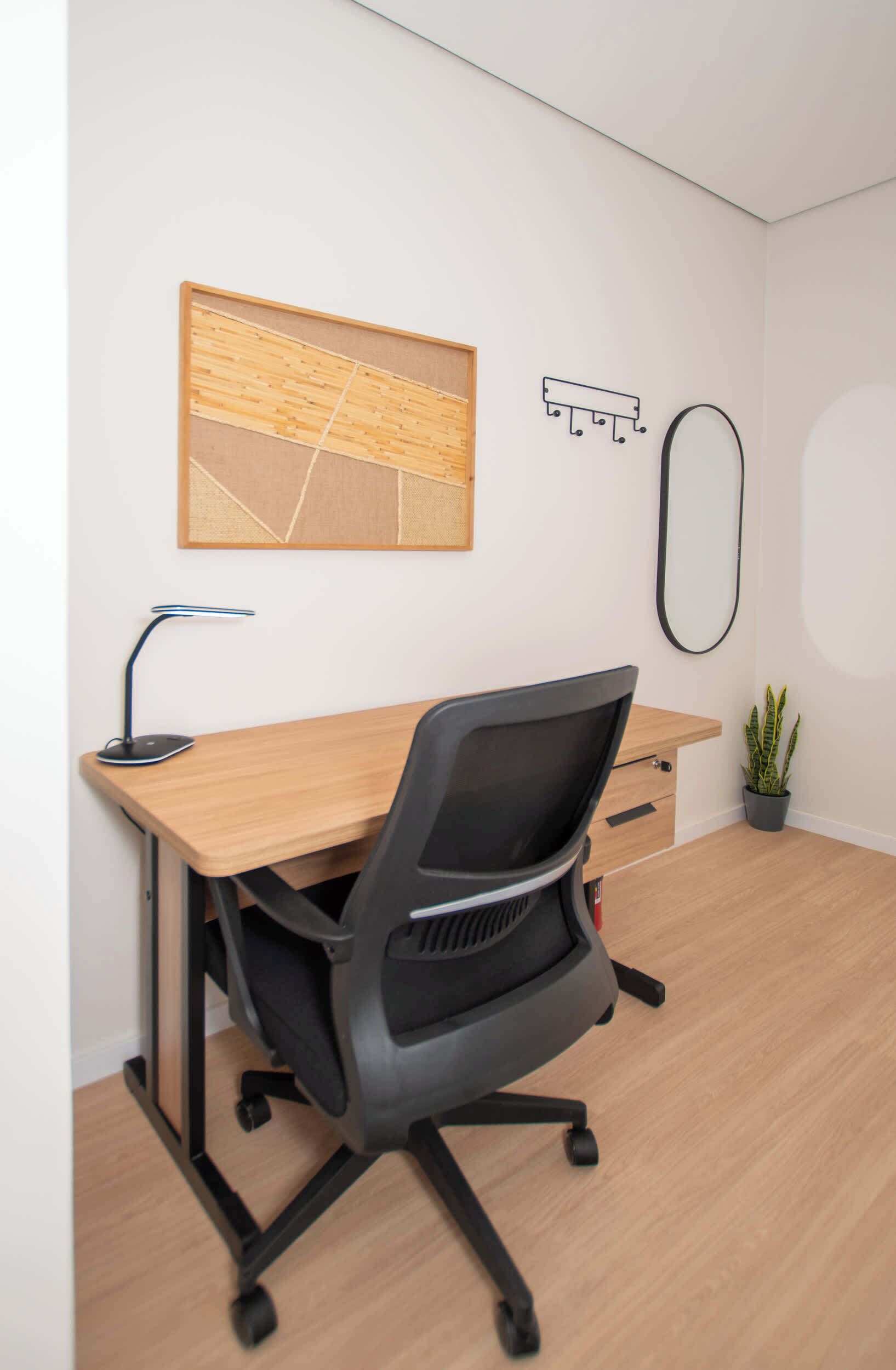 A modern office space with a wooden desk, black office chair, a desk lamp, a framed abstract artwork, a coat hook, a mirror, and a potted plant.