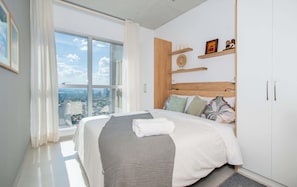 A modern bedroom with a large window, a bed with a white and gray blanket, wooden headboard, and a city view. - Corporate Stays Petra Tower (Asunción)