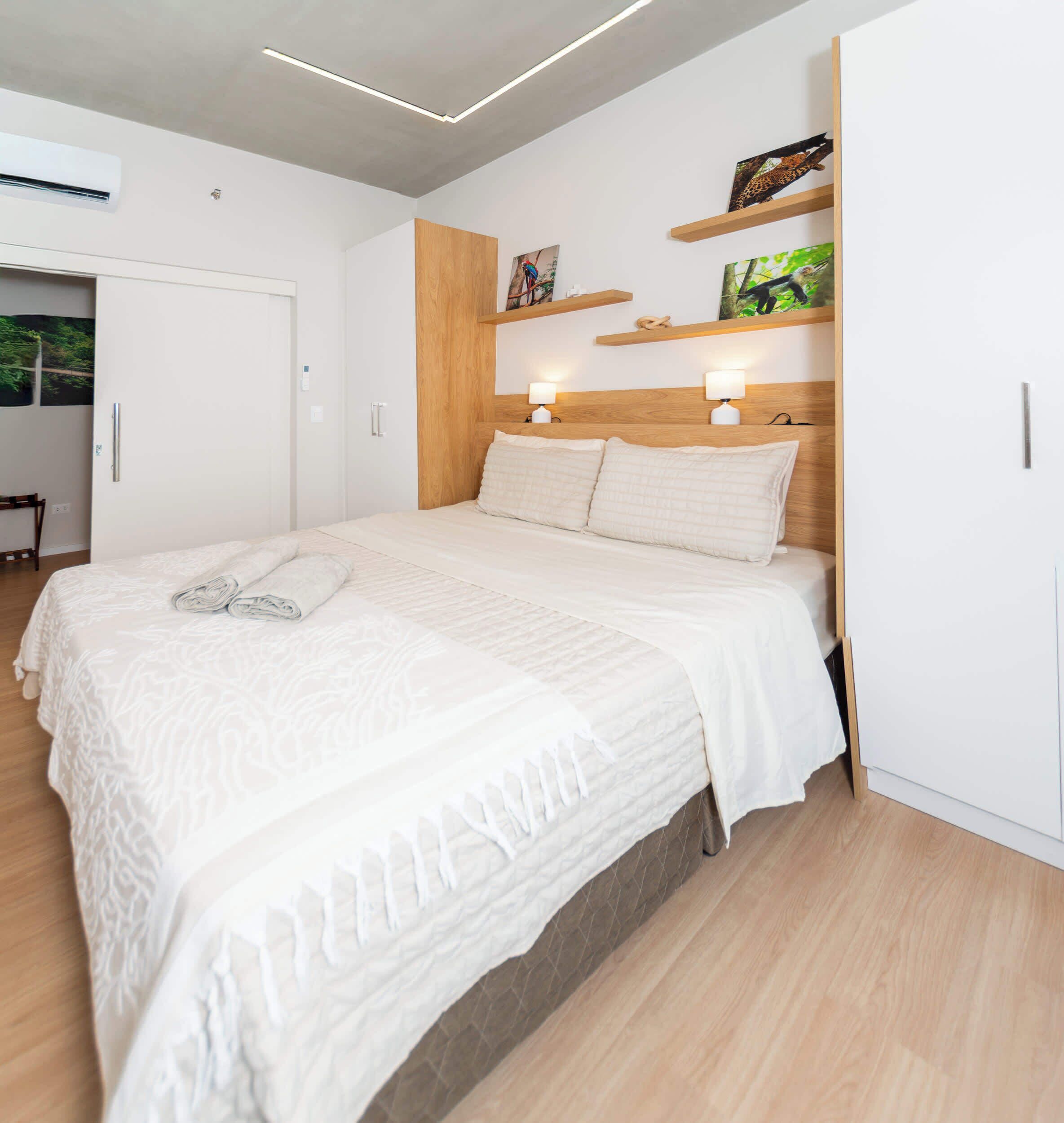 A modern bedroom with a large bed, wooden headboard, wall-mounted shelves with artwork, and a door to a bathroom.