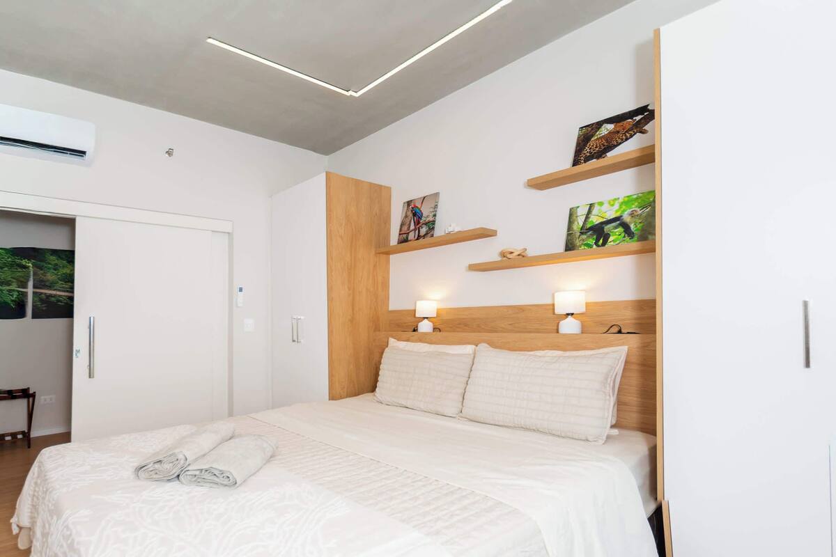 A modern bedroom with a large bed, wooden headboard, wall-mounted shelves with artwork, and a door to a bathroom.