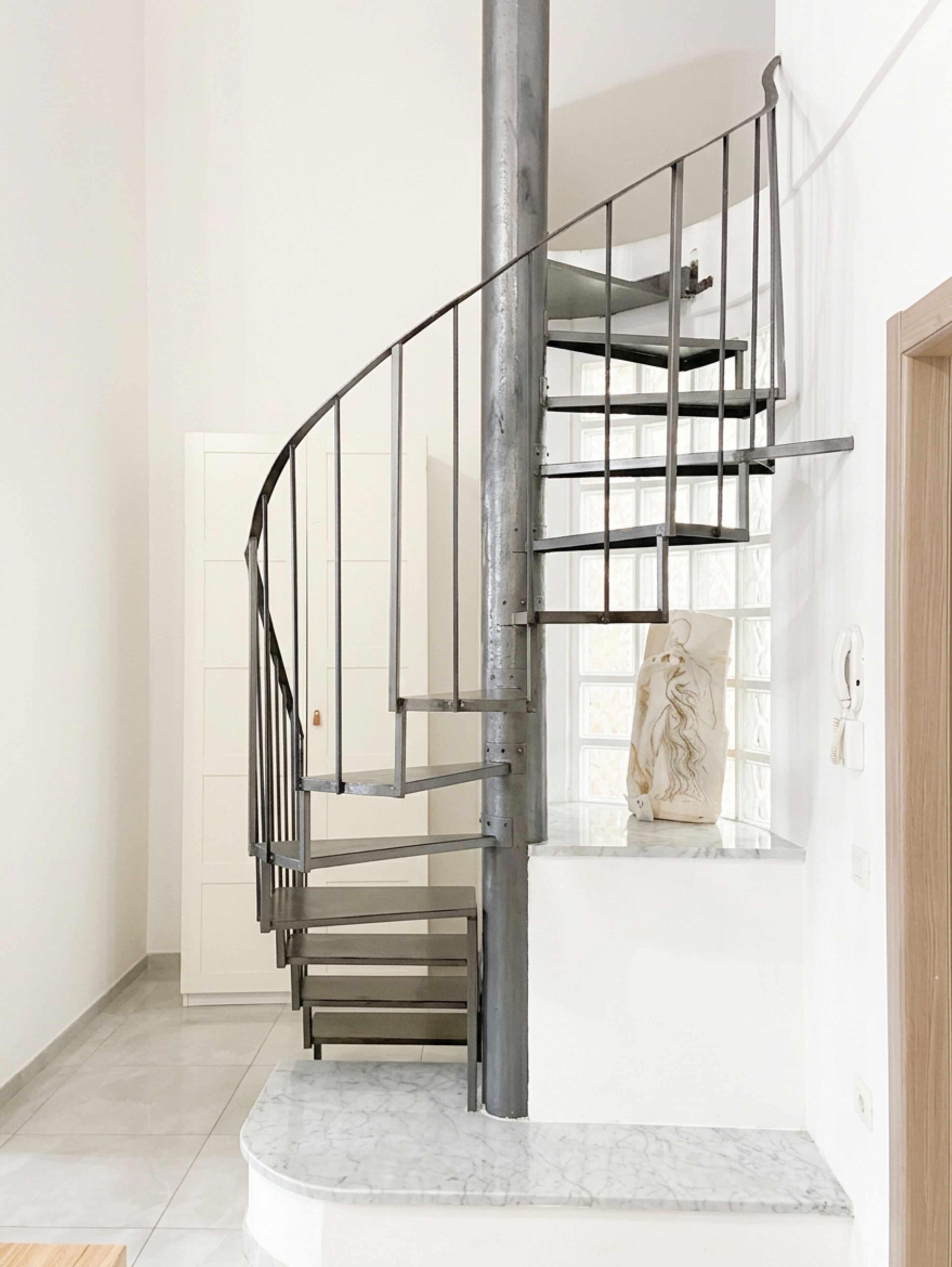 Un escalier en colimaçon moderne, doté de rampes en métal et d’une base en marbre, menant à un palier agrémenté d’un élément décoratif en pierre.