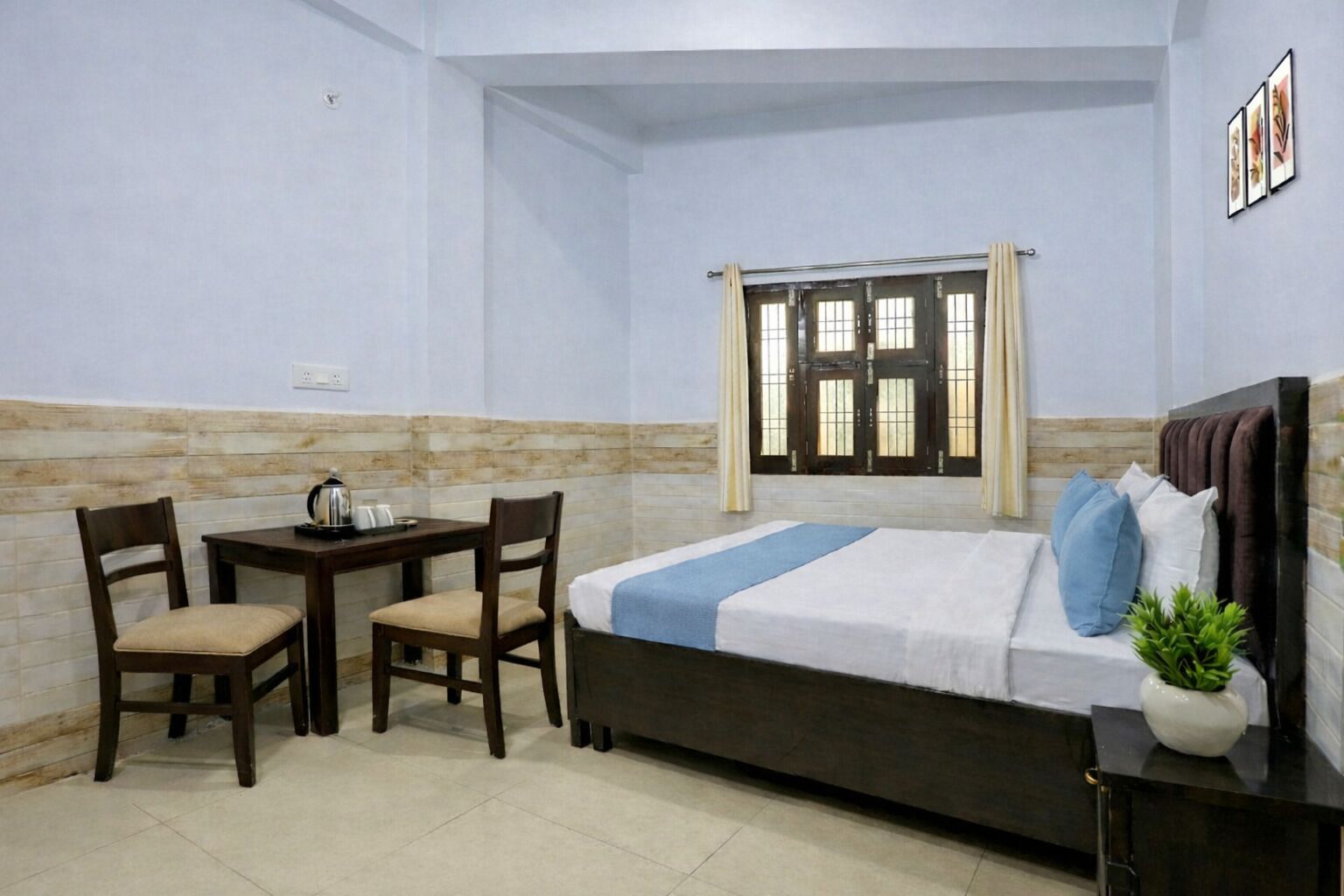 Une chambre à coucher avec un lit, deux chaises, une petite table, une fenêtre avec des rideaux et une plante sur une table de chevet.