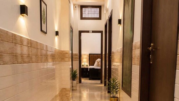 Um corredor de hotel com piso de mármore, plantas em vasos, tapete geométrico e uma cama visível ao fundo.