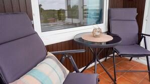 En balkong med två stolar, ett litet bord och utsikt över en byggnad genom ett fönster.