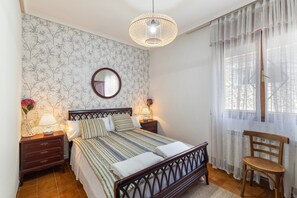 A bedroom with a bed, bedside tables, a mirror, a hanging light fixture, a chair, and a window with sheer curtains.