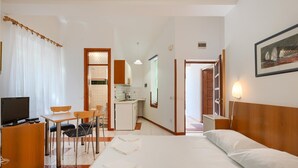 Quarto de hotel com cama, escrivaninha com cadeira, pequena cozinha e banheiro visível por uma porta aberta.