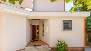 Entrada de um edifício com caminho de azulejos, porta, janela com persianas e um pequeno jardim.