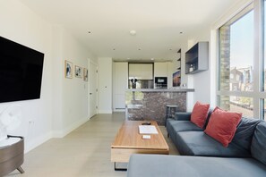 A modern living room with a flat-screen TV, a wooden coffee table, a grey sofa with red cushions, and a kitchen area in the background. - Modern Parkside Flat with Balcony in Kilburn (Greater London)