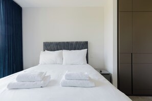 A neatly made bed with white linens and towels, a grey upholstered headboard, and a small bedside table with a lamp. - Modern Parkside Flat with Balcony in Kilburn (Greater London)