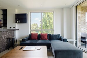 A modern living room with a sectional sofa, a wooden coffee table, and a large window offering a view of trees. - Modern Parkside Flat with Balcony in Kilburn (Greater London)