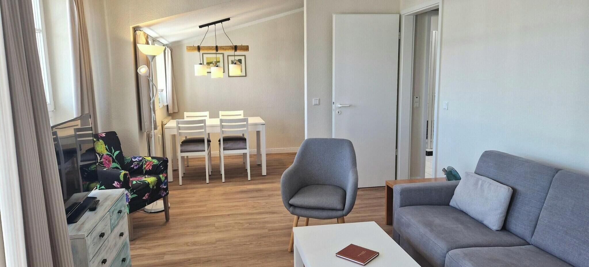 A modern living room with a grey sofa, a floral armchair, a dining area with a table and chairs, and a hallway leading to a door.