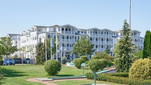 Um edifício grande, com vários andares, fachada simétrica, múltiplas varandas e um jardim bem cuidado com um pequeno espaço de recreação.