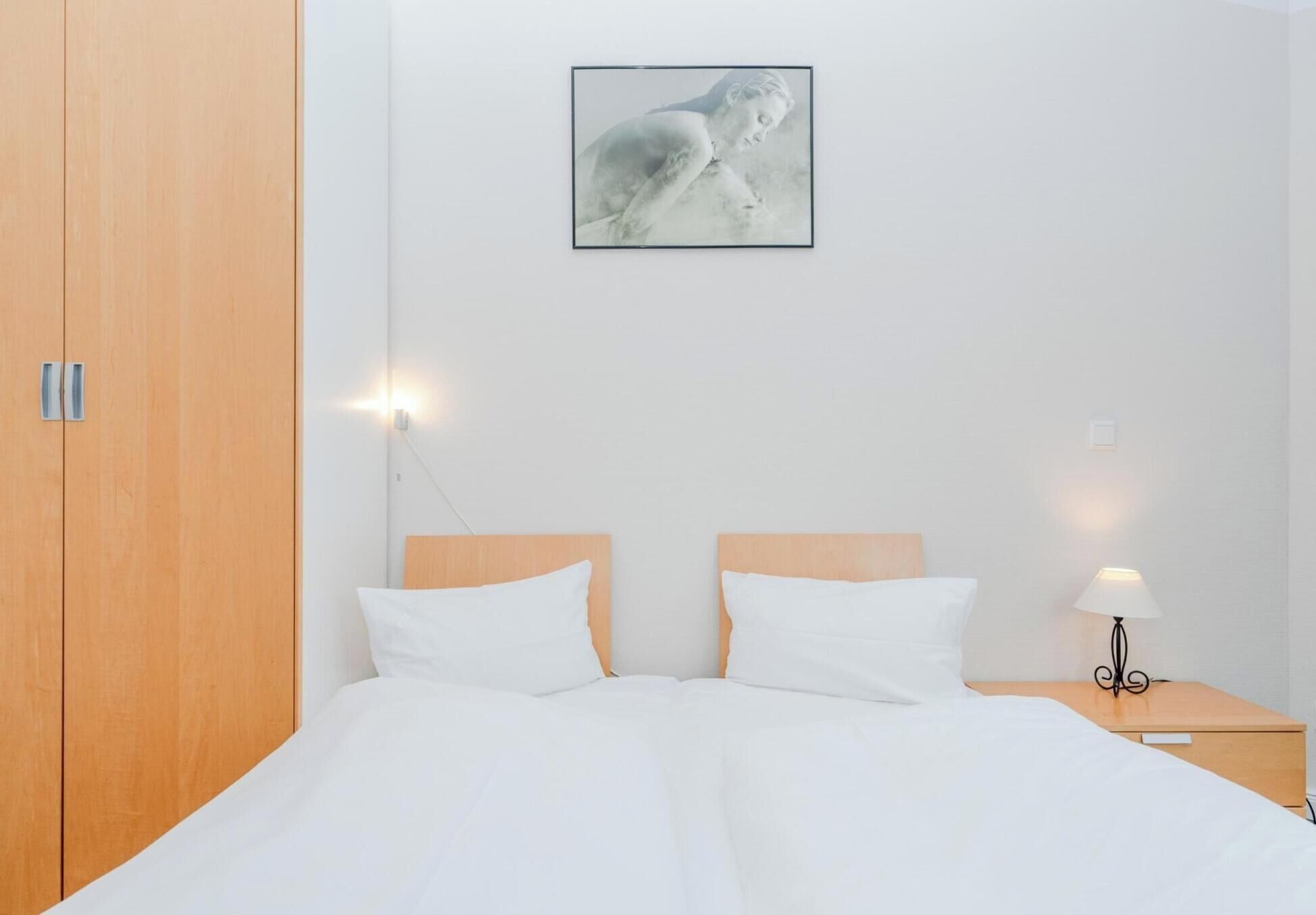 A bedroom with a wooden wardrobe, a bed with white bedding, two bedside tables with lamps, and a framed picture on the wall.
