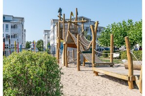 A playground with a wooden climbing structure and a slide, surrounded by sand and greenery. - Ostseeurlaub in der Ferienwohnung Seepark Sellin auf Rügen (Sellin)