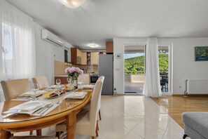 Un comedor con una mesa de madera dispuesta para cuatro, una cocina al fondo y vistas a un paisaje verde a través de una puerta abierta.