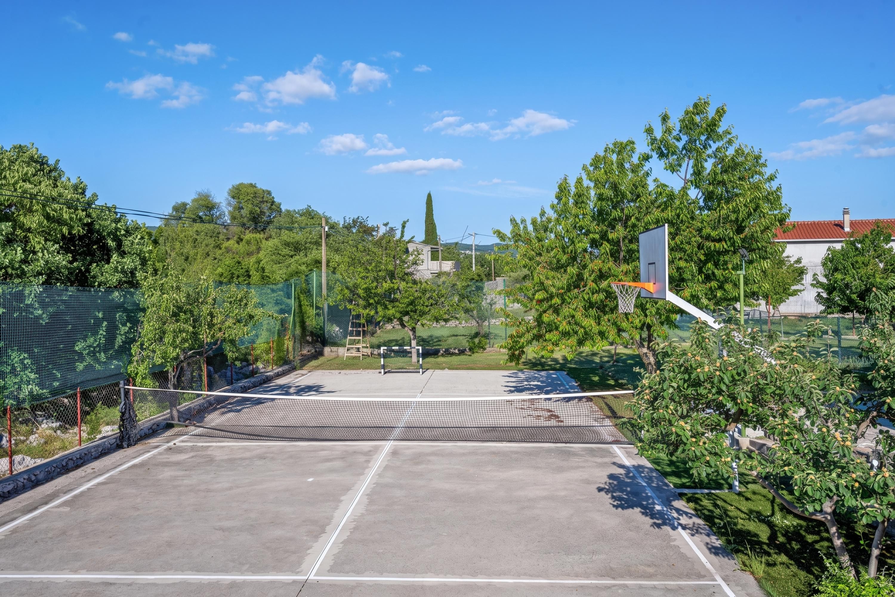 An outdoor basketball court with a net, surrounded by trees and a fence.