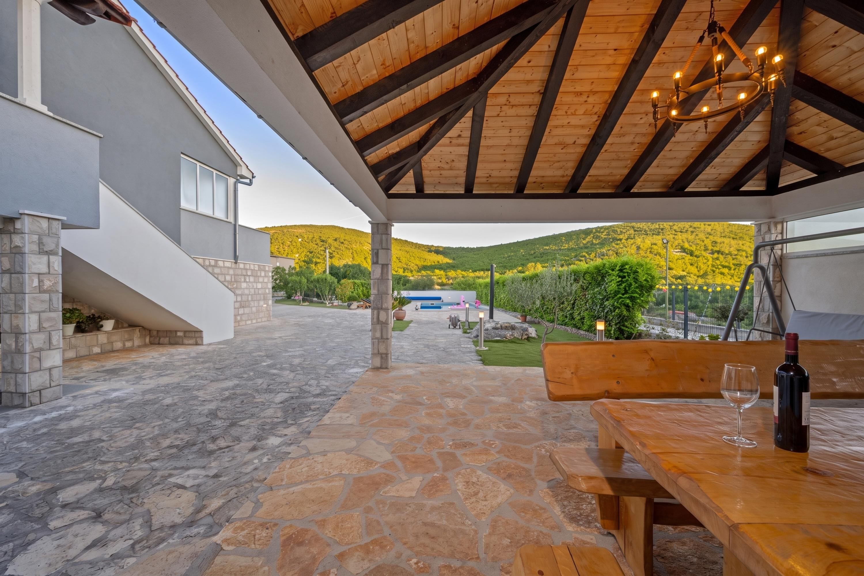 A covered outdoor area with stone flooring, a wooden bench, a table with a wine bottle and glass, and a view of a hillside.