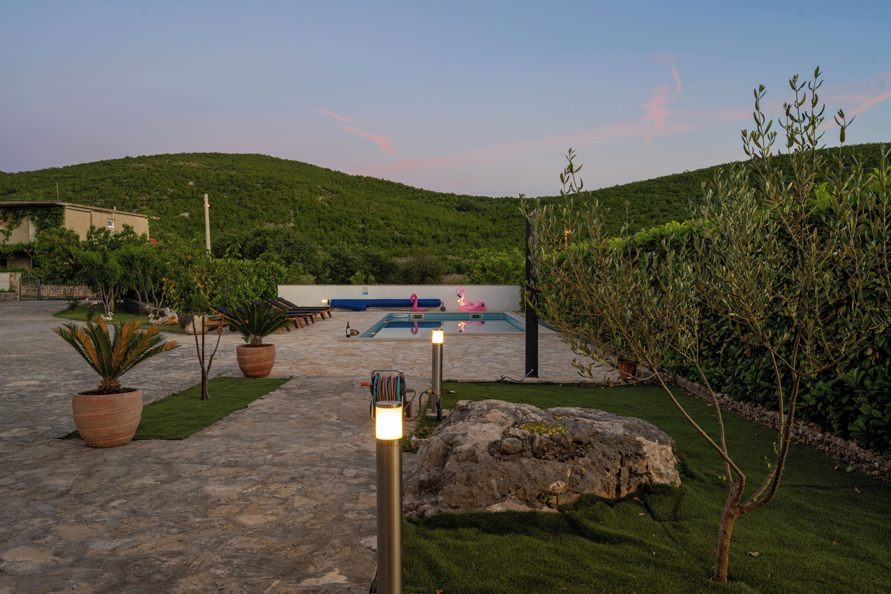 A paved pathway leading to a pool area with potted plants and a rock feature.