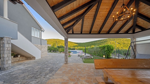 A covered outdoor area with stone flooring, a wooden bench, and a view of a house with a stone staircase. - Pool Villa Marija (Unesic)