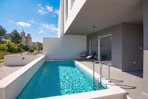 A modern outdoor swimming pool with clear blue water, surrounded by a white deck and a glass door leading to a covered area with chairs. - Pool Villa Pomalo (Vodice)
