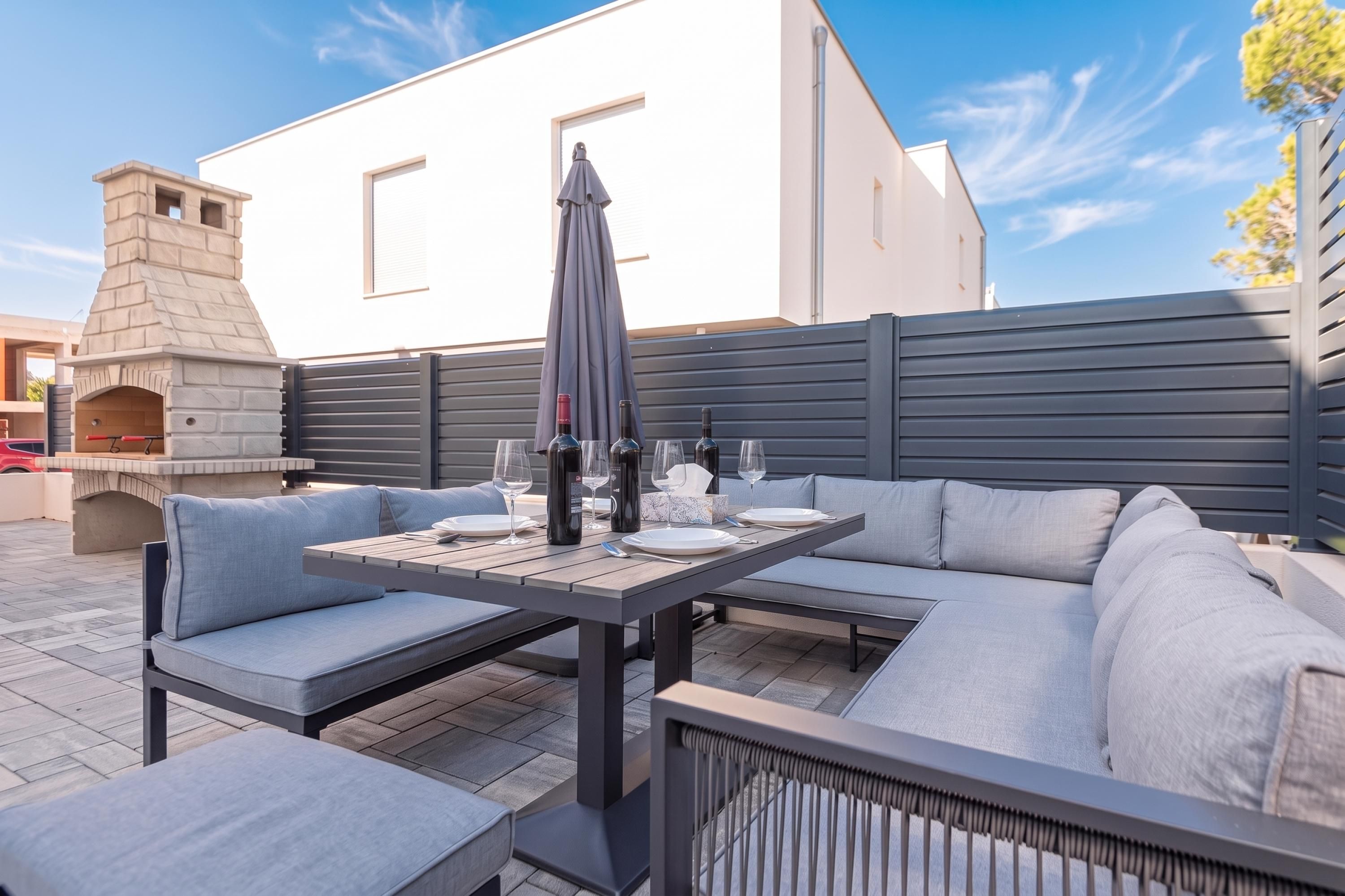 An outdoor patio with a table set for dining, a stone oven, and a modern building in the background.