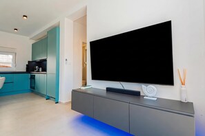 A modern living room with a large flat-screen TV mounted on a grey cabinet, a kitchen area with teal cabinets, and a window with blinds. - Pool Villa Pomalo (Vodice)