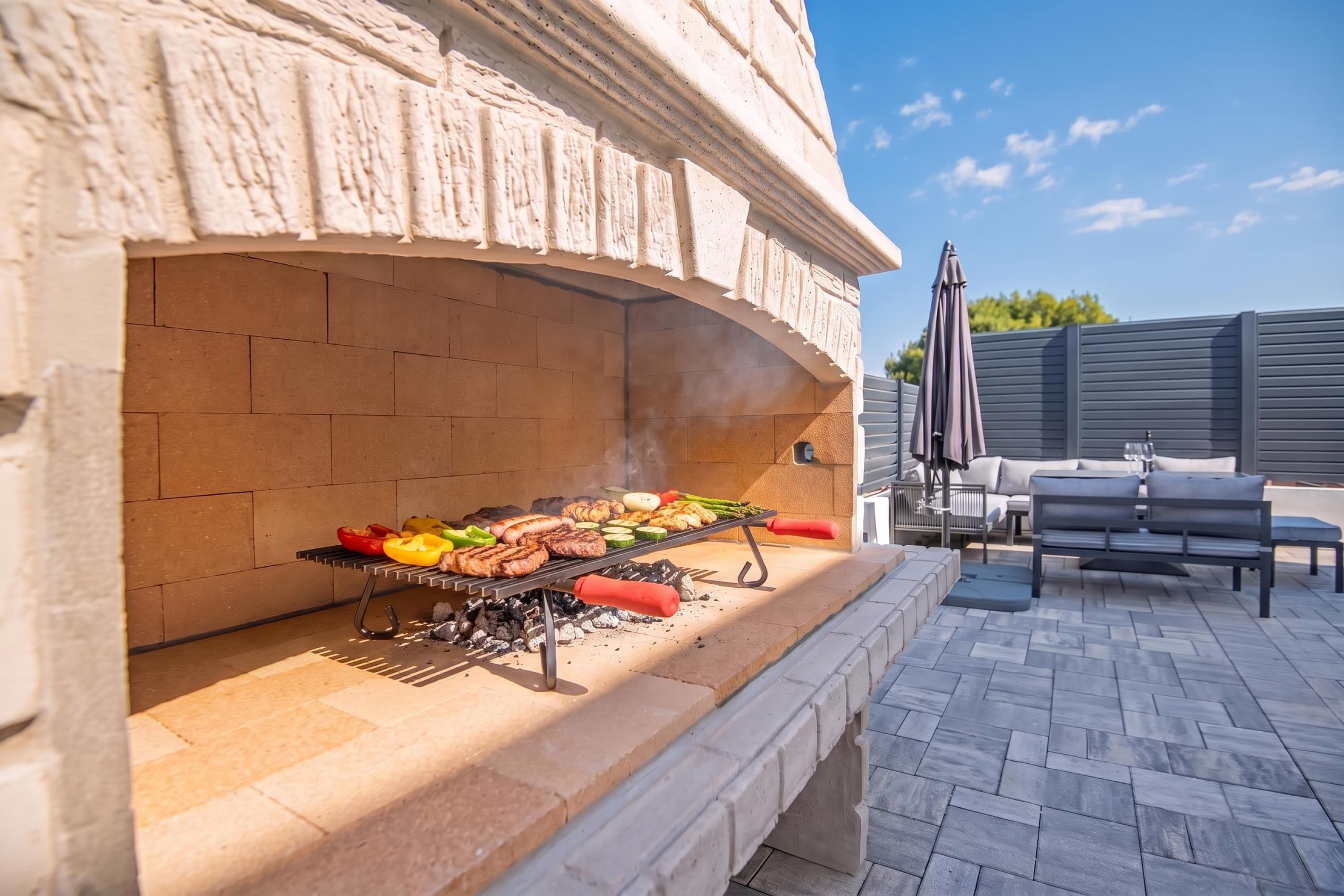 An outdoor grill area with a brick oven, a barbecue grill with various meats and vegetables, and a patio with outdoor furniture.