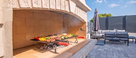 An outdoor grill area with a brick oven, a barbecue grill with various meats and vegetables, and a patio with outdoor furniture.