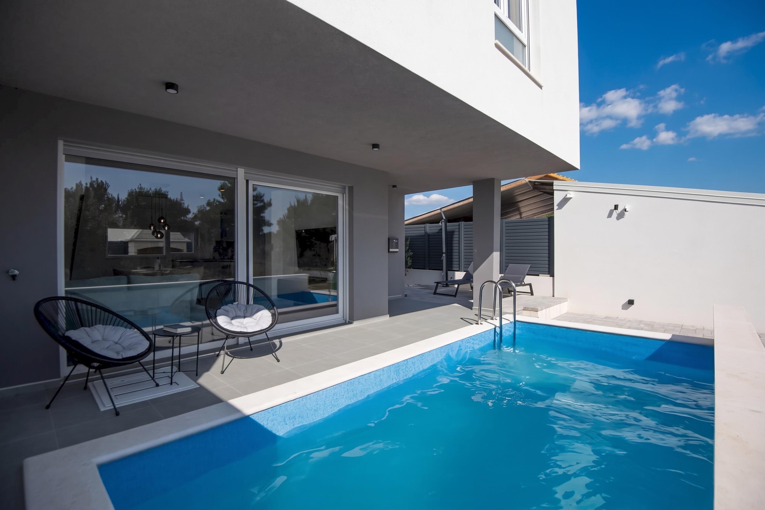 A modern outdoor pool area with glass doors, two lounge chairs, and a clear blue sky.