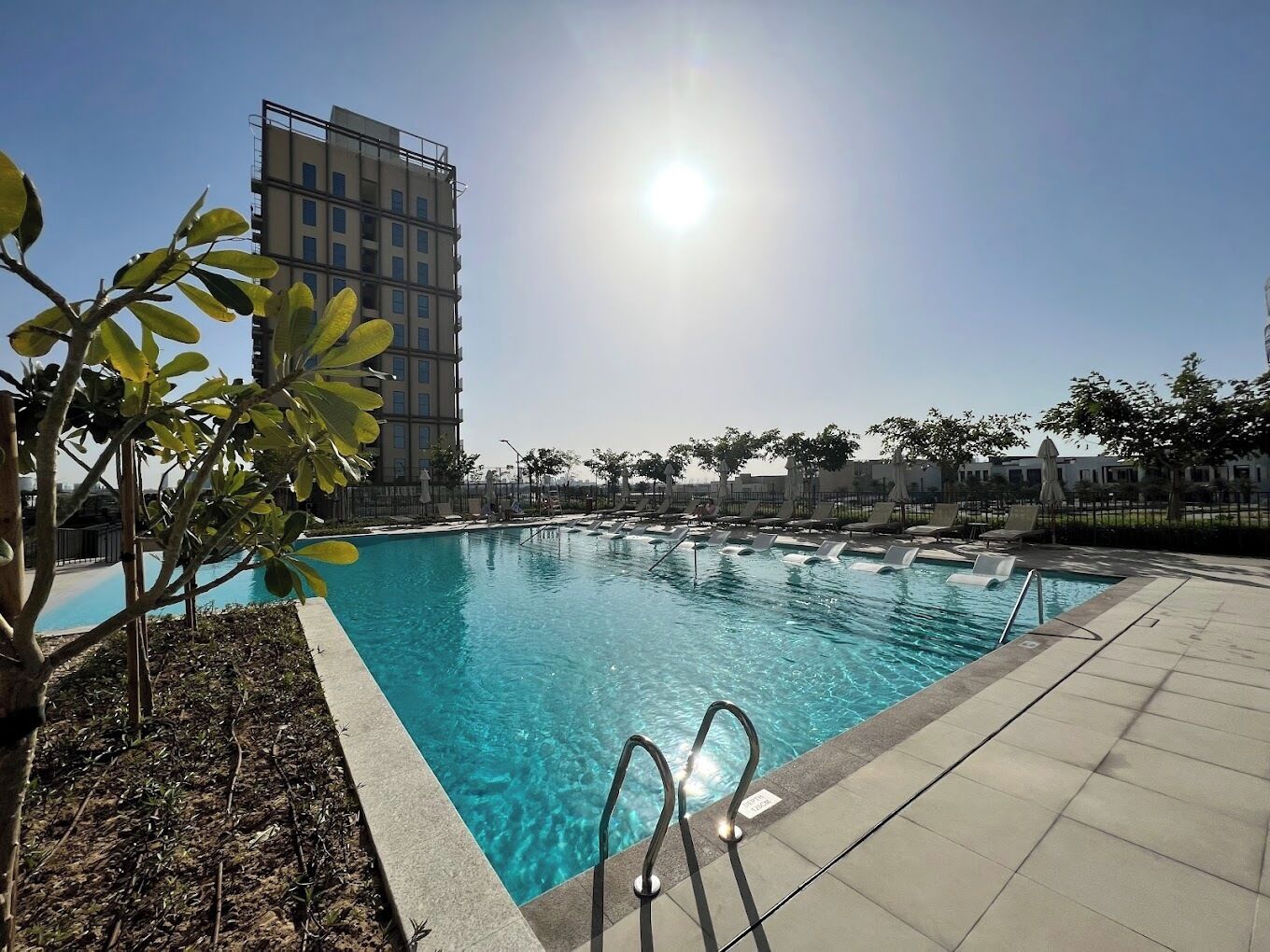 A swimming pool with clear blue water, surrounded by lounge chairs and a tall building in the background.