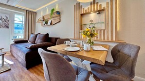 Een moderne woonkamer met een bank, eettafel en stoelen. Een boekenkast met boeken en decoratieve objecten, plus een raam met gordijnen.