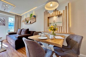 A modern living room with a sofa, dining table, and chairs. There is a bookshelf with books and decorative items, and a window with curtains. - Ferienwohnung in Sellin mit Balkon in Südostlage (Sellin)