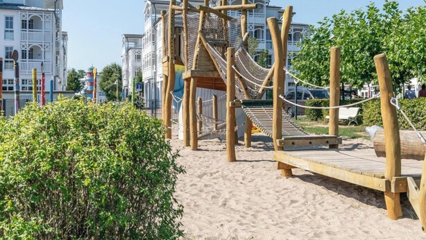 Une structure de jeux en bois, dotée d’un pont de corde et d’un toboggan, entourée de sable et de verdure.