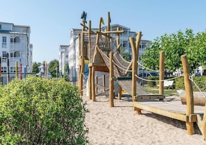 A wooden playground structure with a rope bridge and a slide, surrounded by sand and greenery. - Seepark Sellin Fewo für 3 Personen (Sellin)